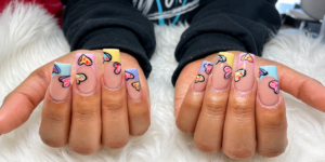 Close-up of pastel French tip acrylic nails with colorful hand-painted heart nail art on a nude base, featuring square-shaped nails in a glossy finish.