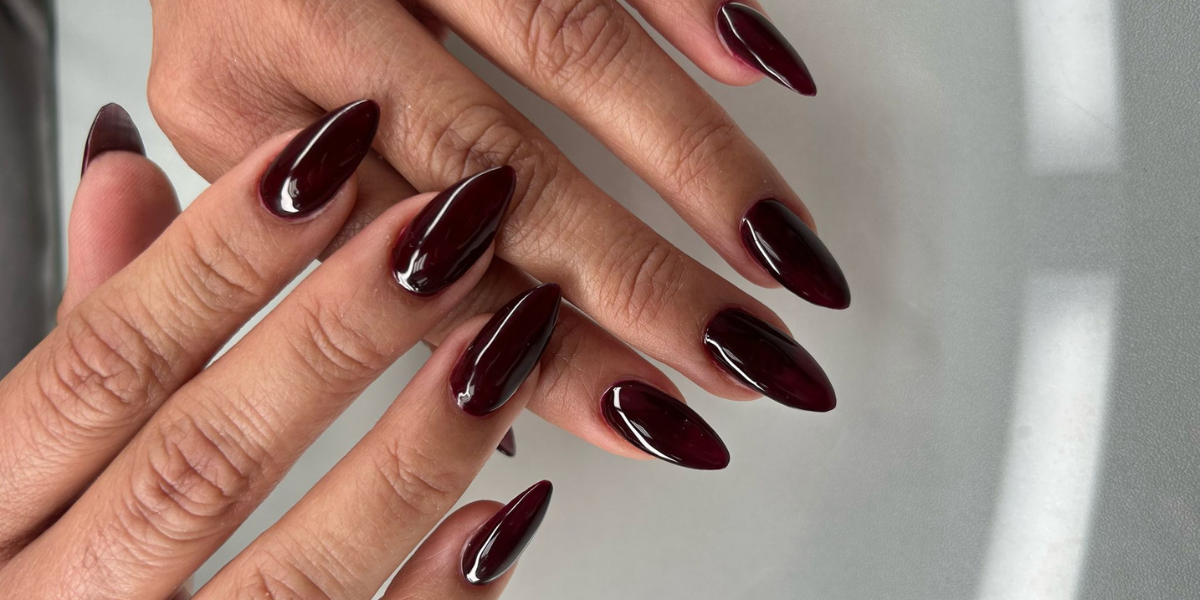 Close-up of hands with long almond-shaped nails featuring a glossy deep burgundy manicure. The rich wine-colored polish has a high-shine finish, highlighting the elegant nail shape against a soft neutral background.