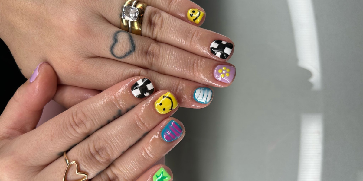 Close-up of hands with short square-shaped nails featuring colorful abstract nail art. The manicure includes yellow smiley faces, black-and-white checkerboard patterns, pastel purple floral accents, blue swirl designs, and vibrant pink and green details. The person is wearing gold rings and has a small heart outline tattoo on one finger, with a soft gray background.
