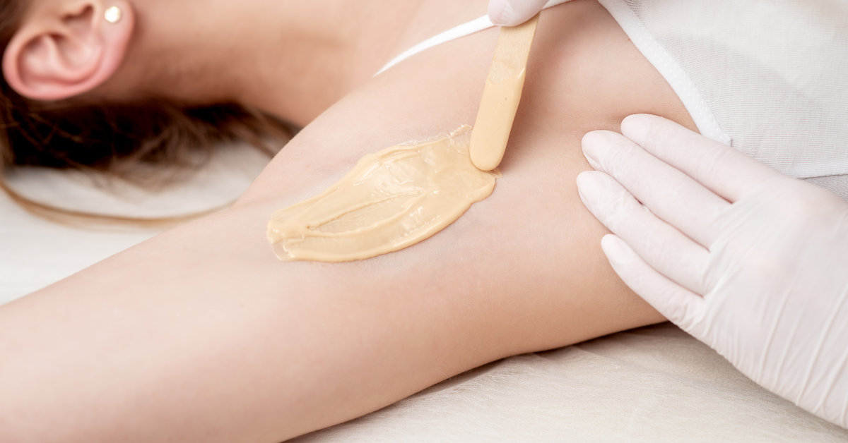 Professional waxing treatment with warm wax being applied to a client’s underarm area using a wooden spatula by a gloved esthetician.