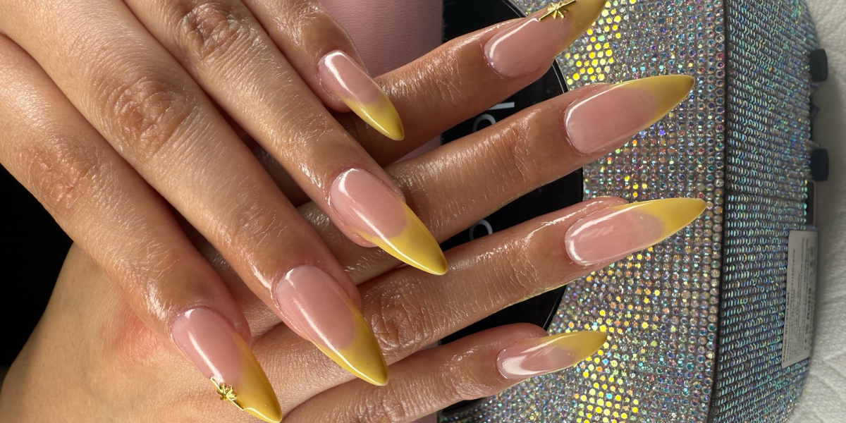 Almond-shaped nude nails with yellow French tips and delicate gold accents at a nail salon in Bellflower.